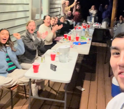Friends cheering on a covered wooden porch at night, gathered around a long folding table with red cups, pitchers and snacks in a selfie-style group photo.