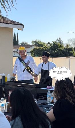 Sunny suburban backyard birthday party with a man wearing a paper crown and sash watching a chef in a black hat and striped apron flip a spatula on a flat-top grill as guests sit at a table with drinks.