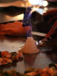 Onion 'volcano' aflame on a teppanyaki griddle, blue flames rising while a spatula hovers over sizzling mixed vegetables.