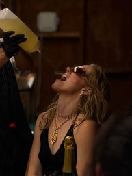 Woman in sunglasses tilts her head back with mouth open as a server pours a yellow drink from a squeeze bottle into her mouth at a dimly lit indoor party, champagne bottle and gold necklaces visible.