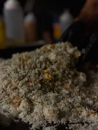 Close-up of a mound of fried rice with diced carrots and scrambled egg on a hot griddle, a gloved hand and condiment bottles blurred in the background