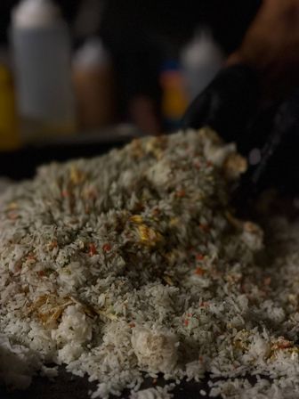 Close-up of a mound of fried rice with diced carrots and scrambled egg on a hot griddle, a gloved hand and condiment bottles blurred in the background