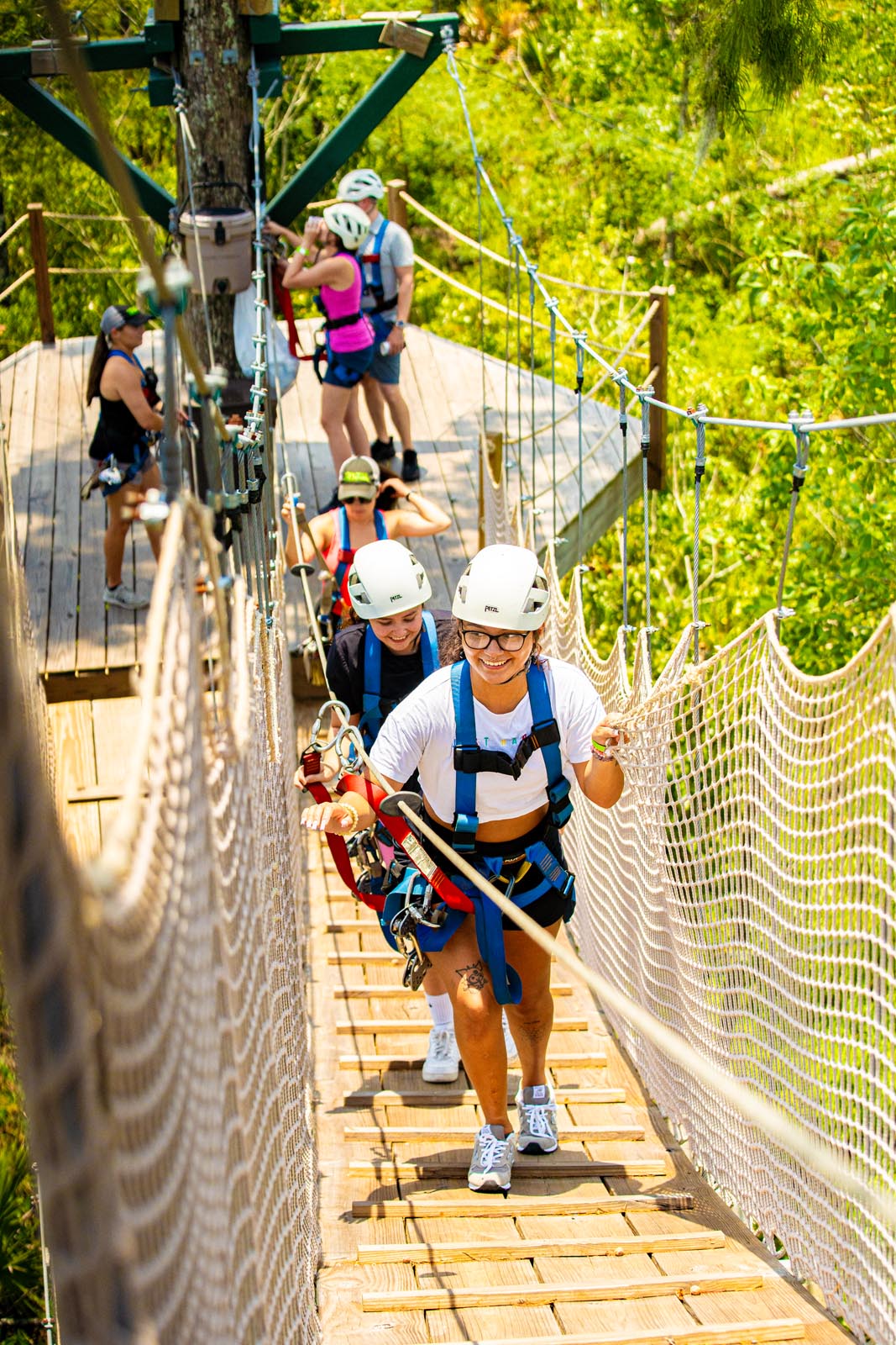 Smiling person in helmet and harness leads a group across a wooden treetop suspension bridge with safety netting on a sunny canopy zipline course in lush green forest.