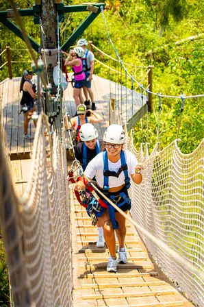 Smiling person in helmet and harness leads a group across a wooden treetop suspension bridge with safety netting on a sunny canopy zipline course in lush green forest.
