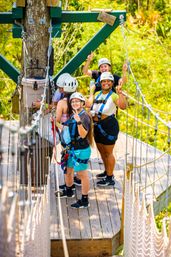 Four people in helmets and harnesses on a wooden zipline platform above a green forest canopy, smiling and flashing peace signs on an outdoor adventure ropes course.