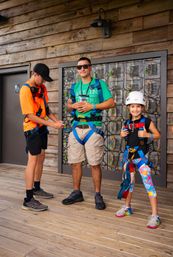 Smiling group suited up for a zipline on a wooden deck — staff in orange adjusts a man’s blue safety harness while a girl in a white helmet and colorful leggings poses in her gear in front of metal lockers.