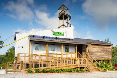 Outdoor adventure building with wooden ramp and deck, corrugated metal siding, spiral-stair zipline observation tower and visible yellow zipline cable against a bright blue sky