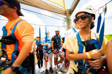 Smiling group in helmets and blue safety harnesses on a wooden platform preparing for an outdoor zipline adventure over green treetops on a sunny day.