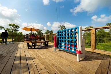 Sunny outdoor wooden deck with oversized blue Connect Four game, picnic table under a yellow umbrella with people seated, surrounded by green trees and blue sky.