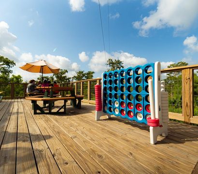 Sunny outdoor wooden deck with oversized blue Connect Four game, picnic table under a yellow umbrella with people seated, surrounded by green trees and blue sky.
