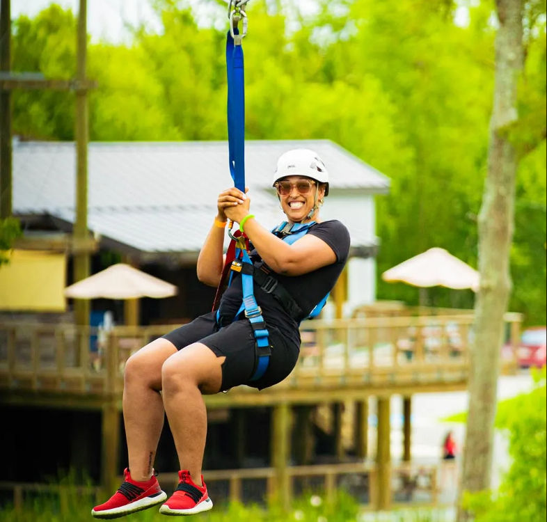 Smiling rider in helmet and harness ziplining above a wooden deck at an outdoor adventure park surrounded by lush green trees