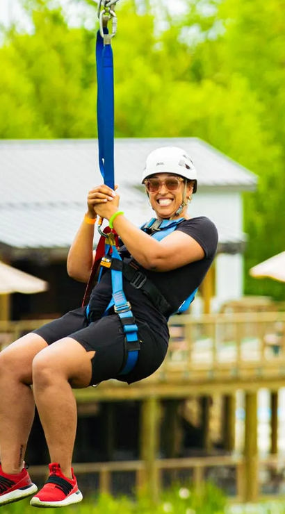 Smiling rider in helmet and harness ziplining above a wooden deck at an outdoor adventure park surrounded by lush green trees