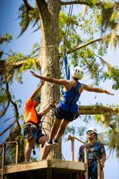 Adventurer in helmet and harness launching from a wooden treetop zipline platform, arms outstretched, assisted by guides against a leafy forest canopy and blue sky.