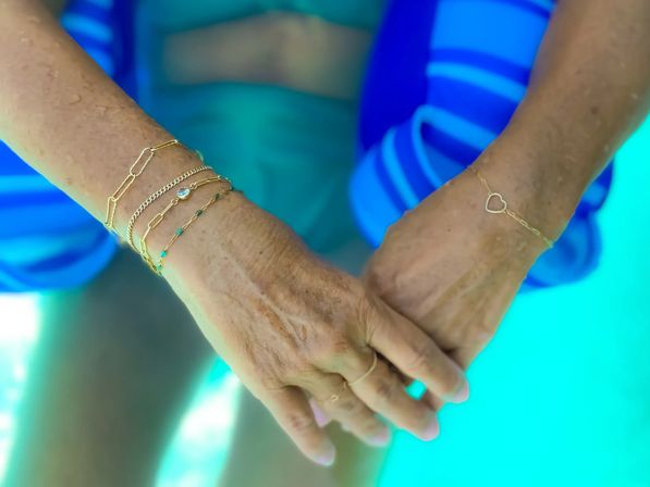 Close-up of two hands clasped poolside over turquoise water, adorned with delicate gold chain bracelets and a small heart charm, blue striped swimwear blurred in the background
