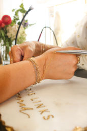 Close-up of an artisan fastening a delicate gold chain bracelet with pliers on a wrist layered with stacked gold bracelets, sunlit marble work surface and blurred flowers in the background.