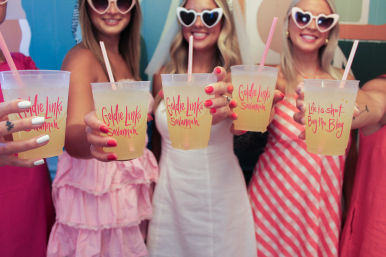 Group of women in pink summer dresses and heart-shaped sunglasses toasting with plastic cups of yellow lemonade with pink logos and straws, festive brunch vibe.