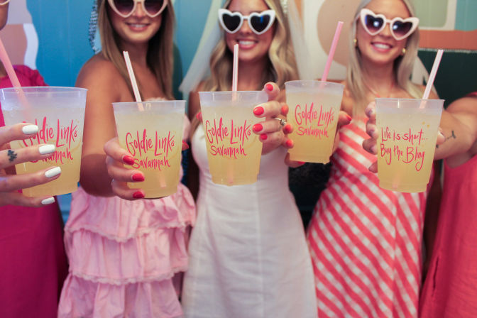 Group of women in pink summer dresses and heart-shaped sunglasses toasting with plastic cups of yellow lemonade with pink logos and straws, festive brunch vibe.
