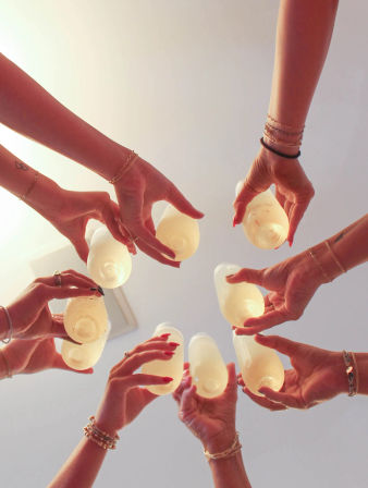 Low-angle shot of a group toast indoors — multiple hands with bracelets and rings holding up plastic cups of pale yellow drinks, festive gathering vibe.