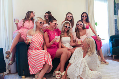 Group of women at a bride-to-be celebration, wearing pink outfits and heart-shaped sunglasses; bride in white veil, everyone laughing and holding drinks on a living room couch.