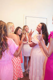 Group of women in pastel pink and white dresses smiling and flashing peace signs while posing in a bright indoor doorway — playful celebration photo