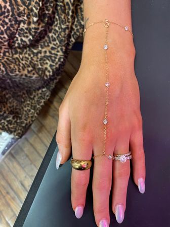 Close-up of a hand wearing a delicate gold hand chain with bezel-set crystals, a chunky gold dome ring, diamond stack rings and iridescent pastel nails over a leopard-print fabric background.