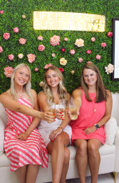 Three smiling women in pink and white dresses clinking champagne flutes on a white sofa against a green floral wall with pink roses and a gold sign that reads Life is short buy the bling