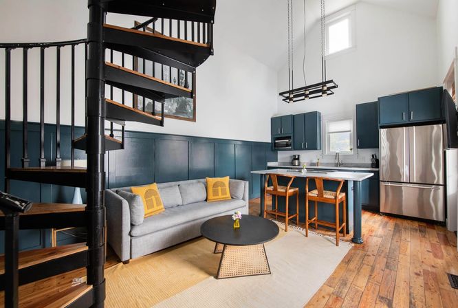 Open-plan loft living room and kitchen with black metal spiral staircase, teal cabinets and stainless fridge, marble-top island with wooden bar stools, gray sofa with yellow accent pillows, round black coffee table and warm wood floors under a high ceiling window.