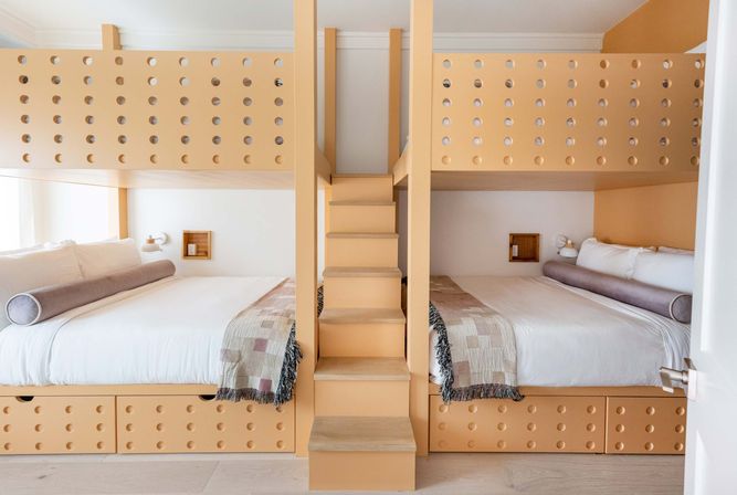 Sunny modern bedroom with peach built-in bunk beds and central stairs, two double lower beds with white linens, gray bolster pillows, patterned throws, and perforated panel details.