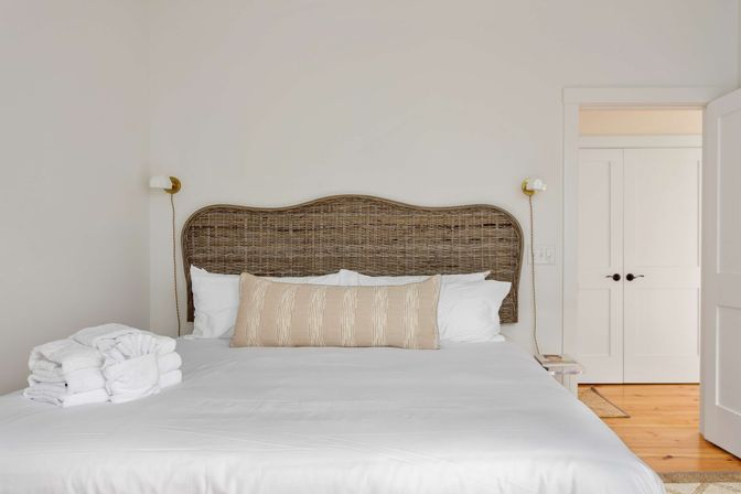 Calm sunlit bedroom with woven rattan headboard, crisp white linens, beige striped lumbar pillow and neatly folded towels on the bed, light wood floor and white closet doors.