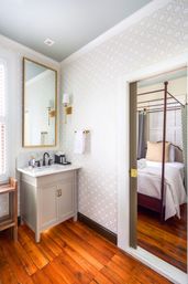 Bright chic ensuite bathroom with marble-topped vanity, gold-framed mirror and brass sconces, geometric wallpaper, warm hardwood floors, open doorway to a four-poster bed in the adjoining bedroom