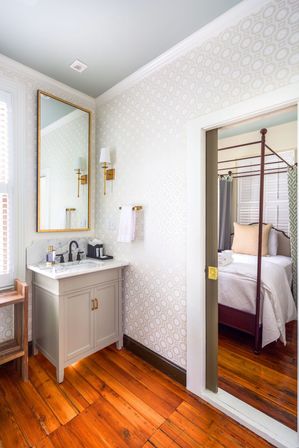Bright chic ensuite bathroom with marble-topped vanity, gold-framed mirror and brass sconces, geometric wallpaper, warm hardwood floors, open doorway to a four-poster bed in the adjoining bedroom