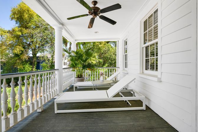Sunny white front porch with decorative balustrade, ceiling fan, two white lounge chairs, small round table with flowers and a potted palm overlooking a tree-lined neighborhood