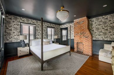 Stylish bedroom with a four-poster bed, botanical wallpaper, dark painted ceiling, exposed red brick fireplace, hardwood floors, neutral area rug, bedside lamps and a tiered chandelier.