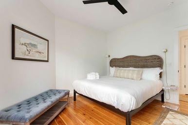 Cozy bright bedroom with a rattan headboard king bed dressed in white linens and a lumbar pillow, folded towels on the bed, tufted gray bench, polished hardwood floors, black ceiling fan, brass wall sconces and framed wall art.