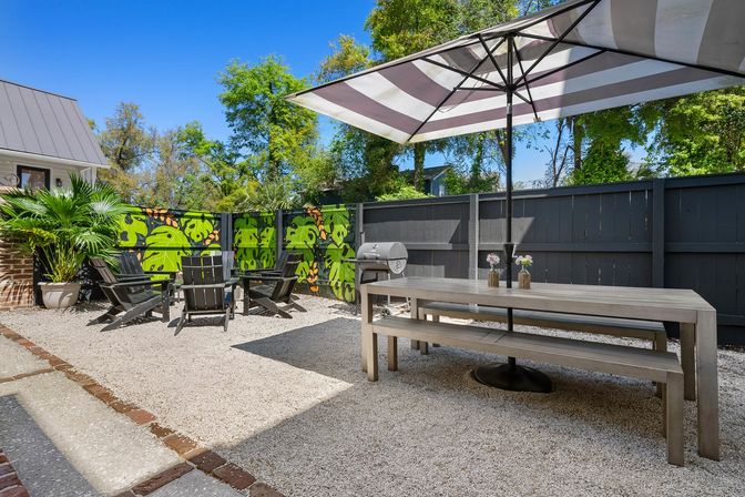 Sunny backyard patio with a long wooden picnic table under a striped umbrella, Adirondack chairs around a small seating area, charcoal grill, potted palms, gravel courtyard and vibrant green leaf mural on a dark privacy fence.