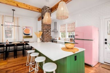 Bright modern farmhouse kitchen with green island and white countertop, pink retro fridge, exposed brick chimney, woven pendant lights, wooden beams and open dining area.