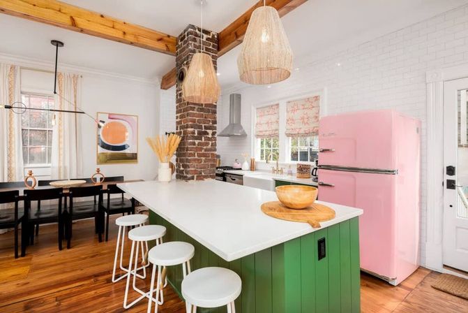 Bright modern farmhouse kitchen with green island and white countertop, pink retro fridge, exposed brick chimney, woven pendant lights, wooden beams and open dining area.