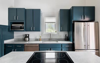 Bright modern kitchen with deep teal cabinets, marble countertops, stainless steel French-door refrigerator, built-in microwave, coffee maker, sink beneath a window, and an island with a black induction cooktop.