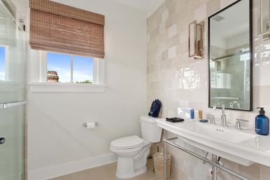 Bright modern bathroom with white sink and chrome fixtures, toilet, glass shower, glossy beige subway tiles, window with woven bamboo shade, blue soap dispenser and toiletries on the counter, and a woven wastebasket.