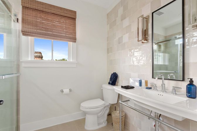 Bright modern bathroom with white sink and chrome fixtures, toilet, glass shower, glossy beige subway tiles, window with woven bamboo shade, blue soap dispenser and toiletries on the counter, and a woven wastebasket.