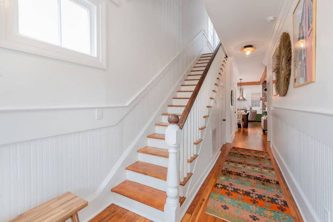 Sunlit modern farmhouse hallway with white beadboard wainscoting, wooden staircase and banister, hardwood floors, and a colorful patterned runner leading toward an open kitchen.