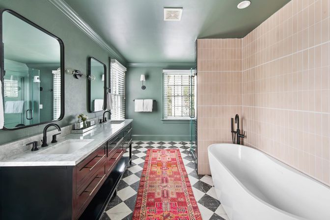 Modern bathroom with sage-green walls and ceiling, double marble vanity with black faucets and mirrors, freestanding white tub by pink tiled shower wall, black-and-white checkered floor and red patterned runner rug.