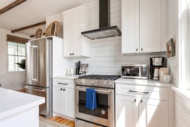 Bright sunlit white farmhouse kitchen with shiplap walls and exposed wood beams, stainless-steel refrigerator and gas range with hood, microwave and coffee maker on quartz countertops, woven shades and hardwood floors.