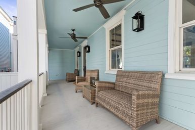 Inviting coastal-style covered porch with light blue siding, two ceiling fans, black wall lanterns and wicker loveseat and chairs with a small side table.