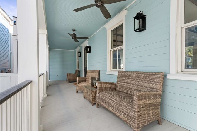 Inviting coastal-style covered porch with light blue siding, two ceiling fans, black wall lanterns and wicker loveseat and chairs with a small side table.