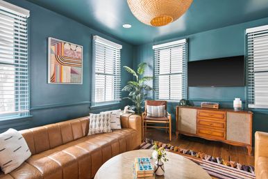 Cozy mid-century modern living room with teal walls, tan leather sectional, wooden credenza and wall-mounted TV, rattan pendant light, potted palm, patterned rug, and coffee table with books and vase.
