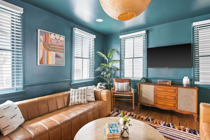 Cozy mid-century modern living room with teal walls, tan leather sectional, wooden credenza and wall-mounted TV, rattan pendant light, potted palm, patterned rug, and coffee table with books and vase.