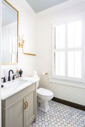 Bright powder room with marble countertop and undermount sink, gray vanity with brass pulls, black faucet, hexagonal marble floor tiles, patterned wallpaper, white plantation shutters and gold wall sconce