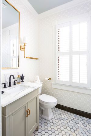 Bright powder room with marble countertop and undermount sink, gray vanity with brass pulls, black faucet, hexagonal marble floor tiles, patterned wallpaper, white plantation shutters and gold wall sconce