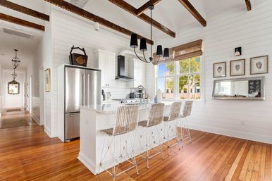 Bright coastal farmhouse kitchen with white shiplap walls, exposed dark wood ceiling beams, hardwood floors, stainless steel refrigerator, white island with four woven bar stools, black pendant chandelier, and a large window with tree views.